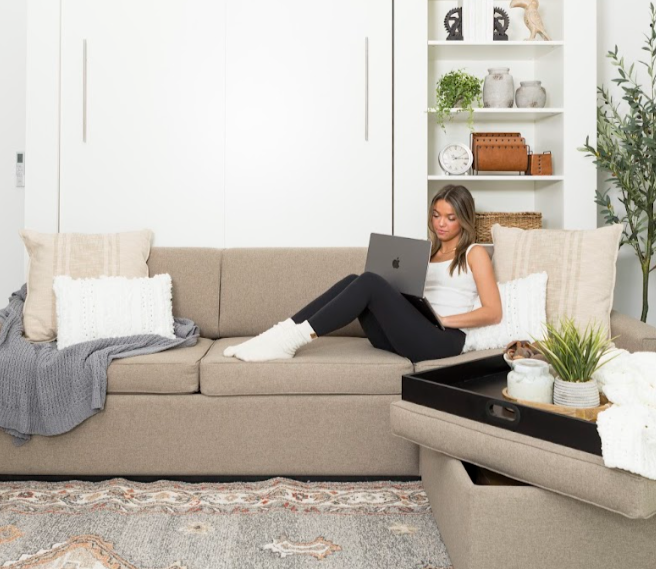 Woman using a laptop on a beige sofa in a living room with a bookshelf and plants.