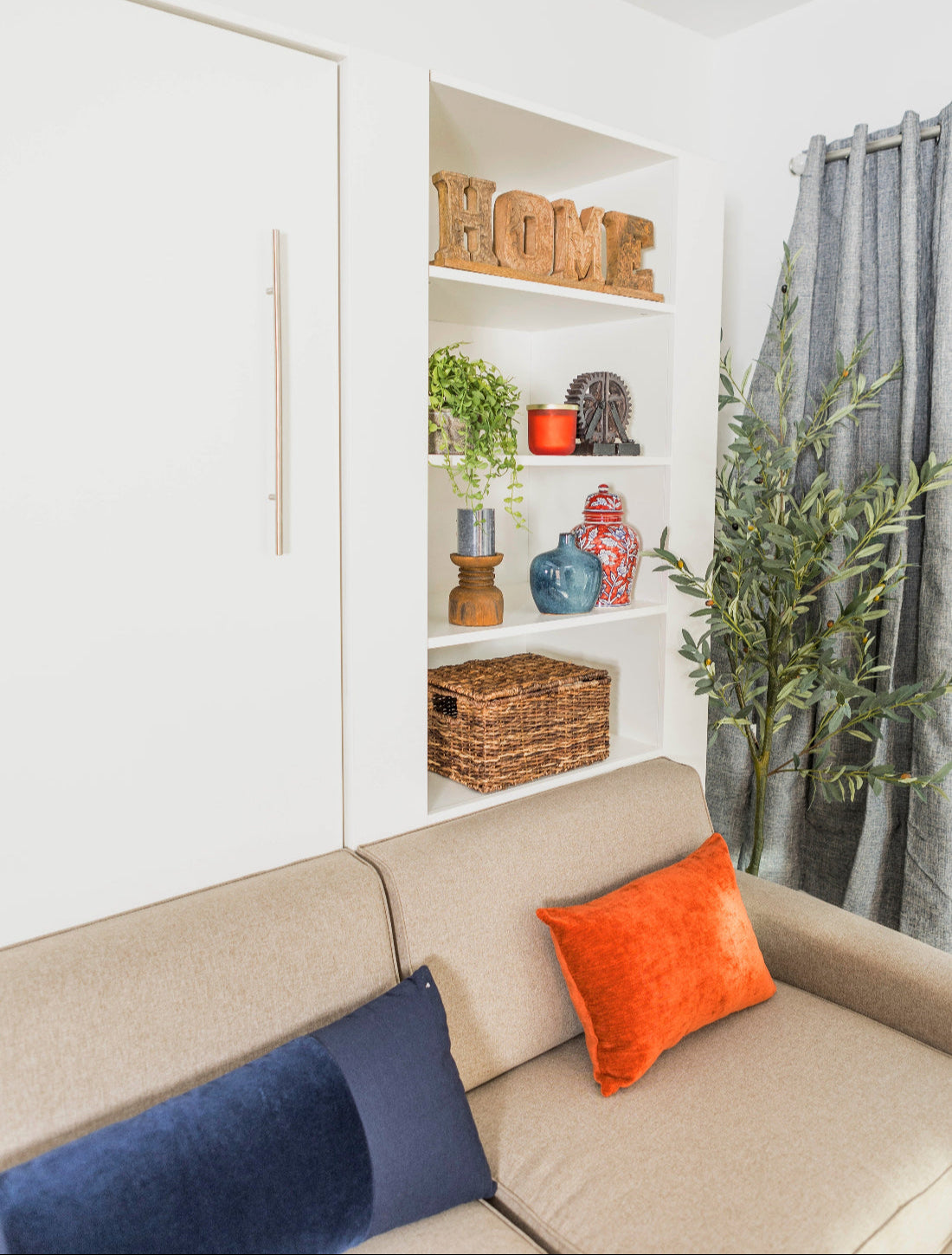 Beige sofa with blue and orange cushions in a room with shelves and a plant.